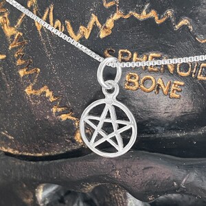 May include: A silver chain necklace with a silver pentagram pendant. The pendant is a five-pointed star with a circle around it. The necklace is on a black background with gold writing.