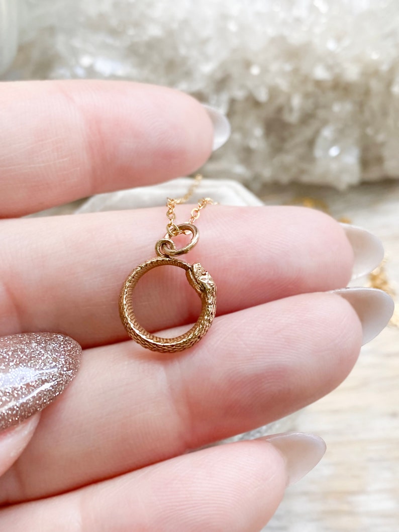 May include: A gold snake necklace with a coiled snake pendant. The snake's head is facing the viewer and its tail is curled around the pendant.