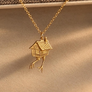 May include: A gold-toned necklace featuring a pendant shaped like a small house with a textured roof, a chimney, and two bird-like legs with talons extending from the bottom. The pendant hangs from a delicate gold-toned chain link necklace.