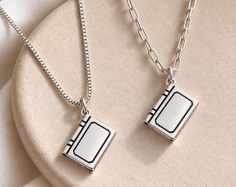 Op de afbeelding: Twee zilverkleurige kettingen met rechthoekige, boekvormige hangers met zwarte gegraveerde omlijningen. De ene ketting heeft een fijne box-schakel, de andere een dikkere, langwerpige schakelketting, beide met een gepolijste metalen afwerking.