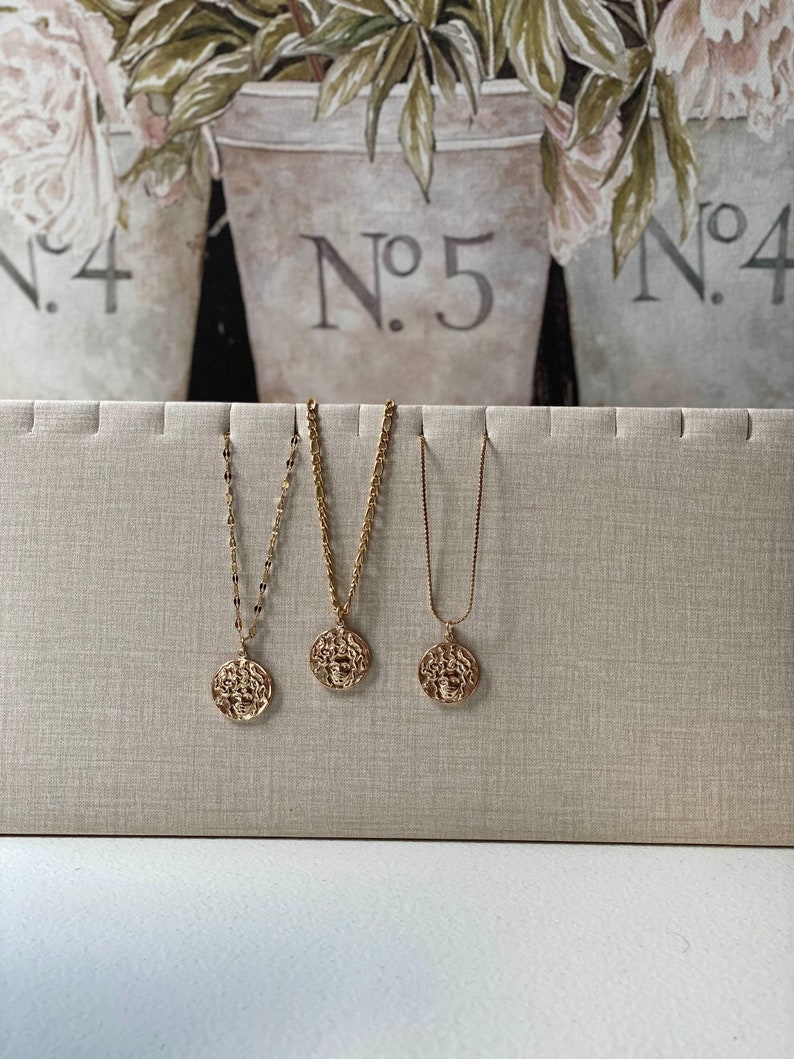 Peut inclure: Trois colliers en or avec des pendentifs ronds pr&eacute;sentant un design d&eacute;taill&eacute;. Les colliers sont pr&eacute;sent&eacute;s sur un fond en tissu beige.