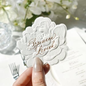 May include: A die-cut white paper place card shaped like a blooming flower with embossed petal details. The name Spencer Levick is printed across the center in elegant, metallic copper-colored cursive script lettering on the textured surface.