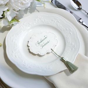 May include: A white scalloped place card with the name Sabrina Turner written in green cursive script. The card features an ornate embossed border and is attached to a light green decorative tassel, resting on a white embossed ceramic plate.