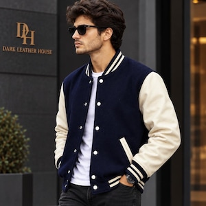 Puede incluir: Una chaqueta universitaria azul marino con mangas color crema, cuello, puños y dobladillo acanalados en blanco con rayas azul marino. La chaqueta cuenta con cierre frontal de botones a presión, dos bolsillos laterales y una camiseta blanca.