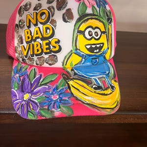 May include: A pink and white trucker hat featuring a hand-painted yellow minion character sitting on a banana. The design includes purple and blue flowers, a leopard print pattern with glitter accents, and the text NO BAD VIBES in yellow letters.