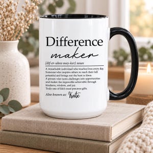 May include: A white ceramic mug with a black interior and handle, featuring black printed text that defines the term Difference maker, followed by a descriptive paragraph and the personalized name Kate at the bottom of the printed text.