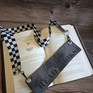 May include: A rectangular dark gray bookmark featuring the text "Pretty Girls READ BOOKS" in a bold, dark font. The bookmark includes silver-toned metal eyelets at each end, attached to a black and white checkered fabric ribbon strap.
