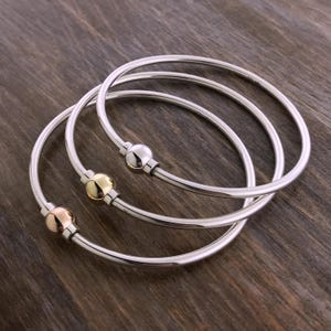 May include: A set of three thin, circular metal bangles. Each bangle features a smooth, polished silver-tone band with a single, small, spherical bead accent. The beads are finished in silver, gold, and rose gold tones respectively.