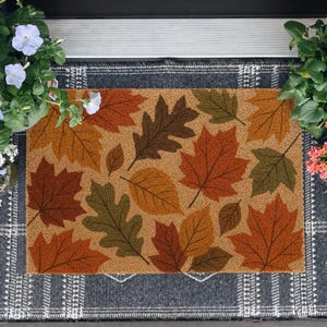 May include: A rectangular doormat featuring a tan background with a pattern of fallen autumn leaves in shades of orange, brown, and olive green. The mat is layered over a dark gray and white plaid rug, flanked by potted blue and pink flowers.