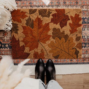 May include: A rectangular coir doormat featuring a printed pattern of various fallen leaves in shades of deep red, burnt orange, brown, and golden yellow. The design displays detailed maple and oak leaf shapes scattered across the natural tan surface.