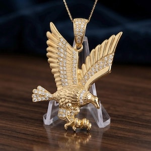 May include: A gold-toned eagle pendant necklace featuring a soaring eagle with wings spread wide. The eagle is encrusted with numerous small, clear, sparkling stones along its wings, body, and tail. It hangs from a gold-toned chain bail.