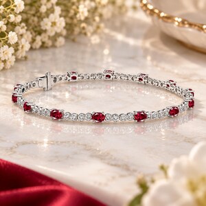 Puede incluir: Una pulsera de metal plateado que presenta una serie de rubíes de color rojo intenso con corte ovalado alternados con pequeñas piedras blancas redondas y transparentes. La pieza incluye un cierre de caja seguro y eslabones circulares.