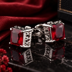 Puede incluir: Un par de gemelos en tono plateado con piedras preciosas de color rojo intenso y corte cuadrado. Los engastes metálicos muestran un intrincado patrón de filigrana calada en los laterales, creando un marco decorativo para las piedras rojas facetadas.