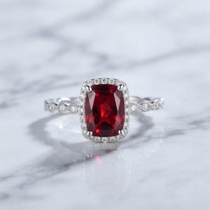Op de afbeelding: Een zilverkleurige ring met een grote, kussengeslepen dieprode edelsteen in het midden. De steen is omringd door een halo van kleine, heldere ronde steentjes. De ringband is aan de zijkanten versierd met een reeks kleine, heldere steentjes.