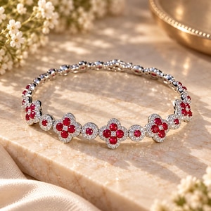 May include: A silver-toned bangle bracelet featuring a repeating pattern of four-petaled flower motifs and circular links. Each flower and circle is set with vibrant red round-cut stones at the center, surrounded by a halo of small white crystals.