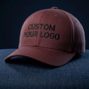 Puede incluir: Una gorra de béisbol de color granate con el texto CUSTOM YOUR LOGO bordado en hilo negro en el panel frontal. La gorra tiene una corona estructurada, una visera curva y ojales cosidos en los paneles superiores, ofreciendo un diseño sencillo.