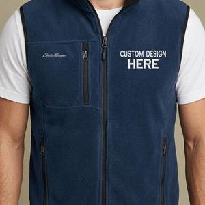 Può includere: Un gilet in pile blu navy con cerniera frontale nera, colletto rialzato e tasche nere con zip sul petto e sui lati. Il logo Eddie Bauer si trova sul petto destro, mentre il testo CUSTOM DESIGN HERE è posizionato sul lato sinistro.