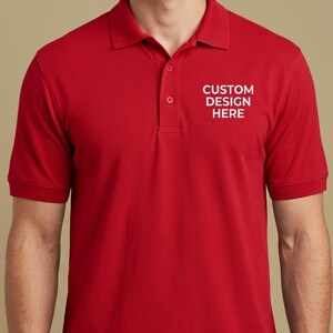 May include: A bright red short-sleeved polo shirt featuring a classic collar and a three-button placket. The left chest area displays the white text CUSTOM DESIGN HERE, printed in a clean, sans-serif font against the solid red fabric background.