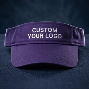 Könnte beinhalten: Ein dunkelvioletter Sonnenschutz mit dem in Weiß gestickten Text CUSTOM YOUR LOGO mittig auf der Vorderseite. Der Sonnenschutz verfügt über einen strukturierten Schirm mit sichtbaren Nähten und ein verstellbares Riemendesign.