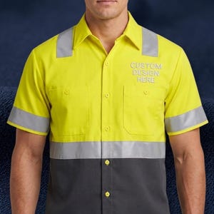 Puede incluir: Una camisa de trabajo de manga corta de alta visibilidad con una sección superior amarilla brillante y una sección inferior gris oscuro. La camisa incluye franjas horizontales plateadas reflectantes en las mangas y el torso, dos bolsillos en el pecho y el texto CUSTOM DESIGN HERE.