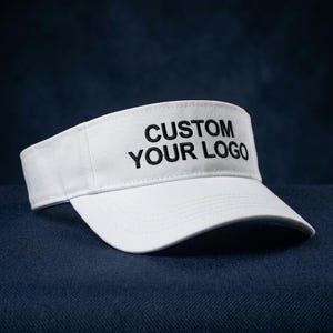 Könnte beinhalten: Ein weißes Visier mit dem schwarz gestickten Text CUSTOM YOUR LOGO auf der Vorderseite. Das Visier hat eine strukturierte Krone und einen gebogenen Schirm, gefertigt aus einem einfarbigen weißen Stoff mit passenden Ziernähten im gesamten Design.