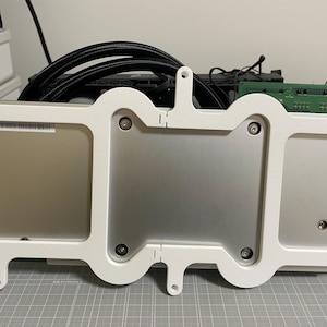 Op de afbeelding: Een witte 3D-geprinte montagebeugel met een gebogen, organisch frameontwerp. De beugel heeft drie rechthoekige uitsparingen, elk met een metalen zilverkleurig onderdeel. Het bevat verschillende kleine ronde schroefgaten langs de randen.