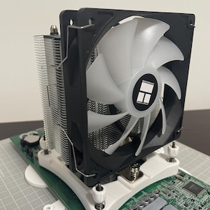 May include: A computer CPU air cooler featuring a silver aluminium fin stack and a black frame fan with translucent white blades. The fan centre displays the Thermalright logo. The unit is mounted on a green circuit board with white plastic brackets.
