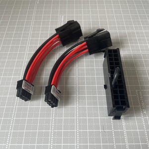 May include: Two black and red electrical power adapter cables with six-pin connectors, each labelled with white stickers reading LEFT and RIGHT, alongside a single black rectangular multi-pin connector block with a small black loop attached to the side.