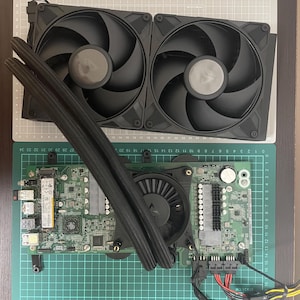 以下が含まれることがあります： 2つの黒いファンを備えた液体冷却ユニット。2本の黒いチューブで緑色の回路基板に接続されており、基板には中央のファン、様々な電子部品、小さな銀色の電池が搭載されています。これらはすべてカッティングマットの上に置かれています。