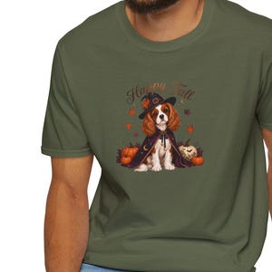 Op de afbeelding: Een olijfgroen T-shirt met korte mouwen en een afbeelding van een Cavalier King Charles-spaniël met een heksenhoed en cape, omringd door pompoenen en herfstbladeren, met de tekst Happy Fall in bruine letters boven de hond gedrukt.