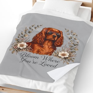 Pode incluir: Uma manta cinza-claro com uma ilustração de um Cavalier King Charles Spaniel castanho rodeado por coroas de flores e o texto Bloom Where You're Loved. A manta está sobre uma estrutura de cama de madeira com almofada e lençóis brancos.