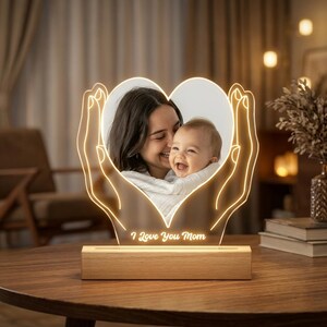 Może przedstawiać: Lampka nocna z akrylu w kształcie serca z nadrukowanym zdjęciem kobiety trzymającej dziecko. Akryl jest podświetlony ciepłym światłem LED w kształcie dwóch dłoni. Podstawa wykonana jest z jasnego drewna z napisem I Love You Mom.