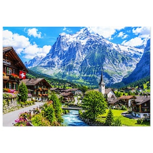 May include: A landscape puzzle featuring a Swiss village with wooden houses, a church with a steeple, and a flowing river. In the background, a massive snow-capped mountain rises against a bright blue sky with white clouds. The scene includes green trees.