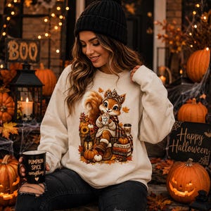 Könnte beinhalten: Ein beiges Sweatshirt mit Rundhalsausschnitt und einem Motiv eines Eichhörnchens mit Leopardenmuster-Stirnband und Schal, das eine Tasse mit der Aufschrift Hello Fall hält, umgeben von Kürbissen, Büchern und Herbstblättern, dazu eine schwarze Mütze.