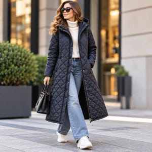 Puede incluir: Un abrigo largo acolchado de color negro con capucha, usado sobre un suéter blanco de cuello alto y jeans azul claro. El abrigo cuenta con cremallera frontal y botones a presión. Se lleva un bolso negro en la mano y zapatillas blancas.
