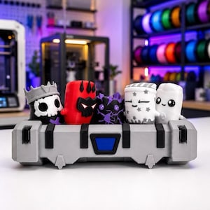 Puede incluir: Un contenedor de almacenamiento rectangular gris que sostiene cinco pequeñas figuras estilizadas. Las figuras incluyen un personaje de calavera con corona, una criatura roja con cuernos, un monstruo de cristal morado, una figura con estrellas y una blanca.