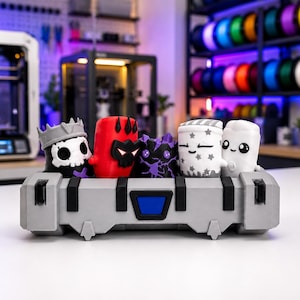 May include: A gray rectangular storage container with black accents holding five small, stylized 3D-printed figures. The figures include a skull character with a crown, a red horned character, a purple crystal creature, and two white figures with star patterns.