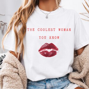 Puede incluir: Una camiseta blanca de manga corta con el texto en rojo THE COOLEST WOMAN YOU KNOW impreso sobre un gráfico de una marca de beso de pintalabios rojo oscuro. La camiseta tiene cuello redondo y es de tela blanca lisa.