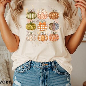 Puede incluir: Una camiseta de color marfil con un estampado gráfico de nueve calabazas dispuestas en cuadrícula. Cada calabaza muestra un patrón único, incluyendo cuadros, estampado de leopardo, lunares y motivos florales, con lazos y follaje otoñal.