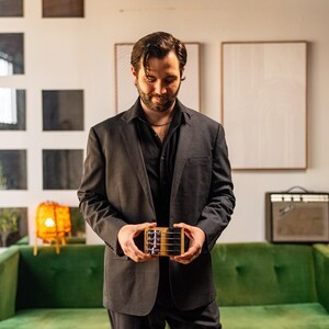 May include: A small, rectangular wooden musical instrument with four metal strings and a bridge, held in both hands. The instrument features a light-colored wood body with a dark top plate and silver tuning pegs visible on the side.