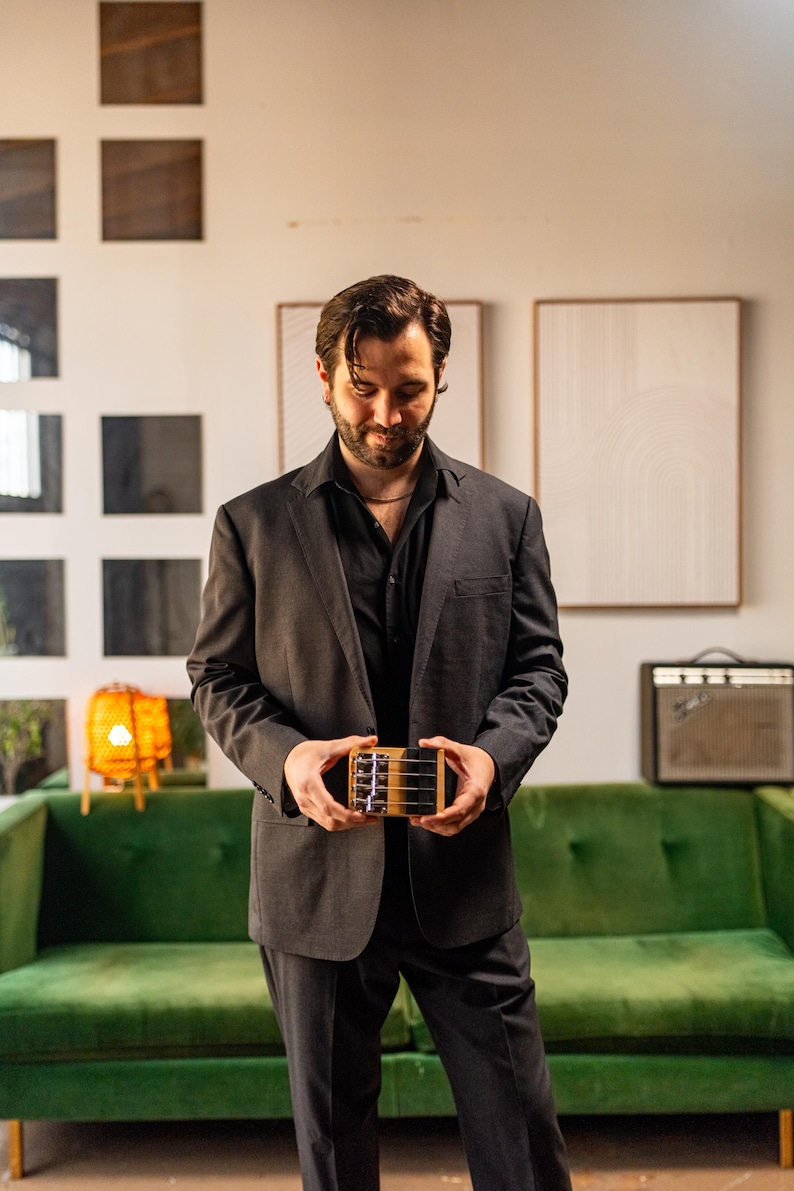 May include: A small, rectangular wooden musical instrument with four metal strings and a bridge, held in both hands. The instrument features a light-colored wood body with a dark top plate and silver tuning pegs visible on the side.