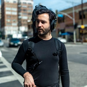 May include: A black long-sleeve shirt worn with a black backpack and black over-ear headphones. The backpack features a black instrument case attached to the back. A small black rectangular device with a red stripe is held in the hand.