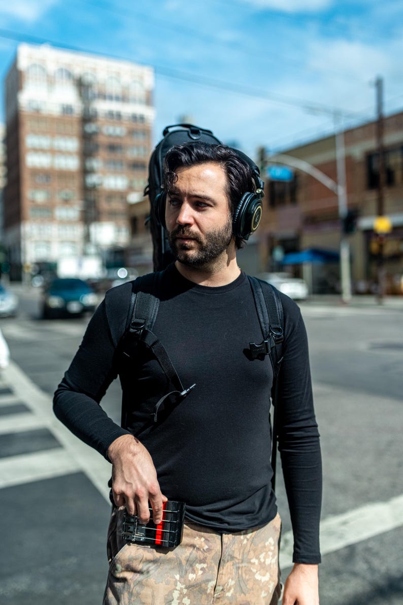 May include: A black long-sleeve shirt worn with a black backpack and black over-ear headphones. The backpack features a black instrument case attached to the back. A small black rectangular device with a red stripe is held in the hand.