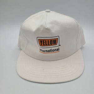 May include: A white baseball cap featuring a rectangular logo on the front panel. The logo contains the word YELLOW in black letters inside an orange box, with the word International written in black below it, underlined by an orange line.