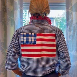 May include: A blue and white gingham button-down shirt featuring a large rectangular American flag patch on the back. The shirt has long sleeves with rolled cuffs and a red and white polka dot fabric detail visible around the collar area.