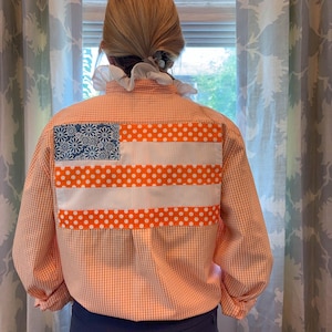 Puede incluir: Una camisa de manga larga con botones en un patrón de cuadros vichy naranja claro y blanco. La parte posterior presenta un parche rectangular con el diseño de una bandera estadounidense, con un cantón floral azul y rayas horizontales de lunares.