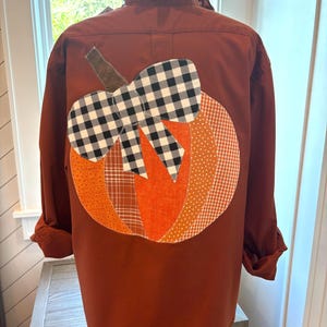 Op de afbeelding: Een roestkleurig overhemd met lange mouwen, knoopsluiting en een ruchekraag. Op de achterkant bevindt zich een groot, geappliceerd pompoenontwerp gemaakt van diverse oranje en bruine stoffen, afgewerkt met een zwart-witte strik met ruitpatroon.