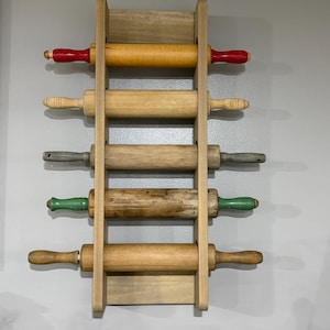 May include: A vertical wooden wall rack holding five rolling pins. The top pin has red handles and a yellow barrel, the second is natural wood, the third is grey, the fourth has green handles, and the bottom pin is natural wood with a dark finish.