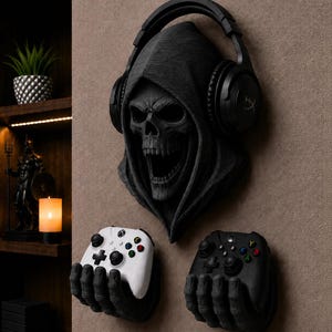 May include: A wall-mounted black skull sculpture wearing black over-ear headphones, positioned above two black skeletal hand wall mounts, each holding a video game controller, one white and one black, with colorful action buttons.
