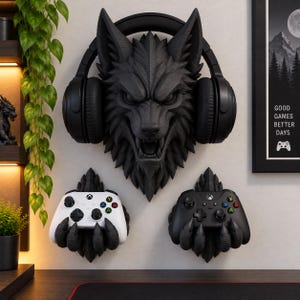 May include: A black wall-mounted wolf head headphone stand holding black over-ear headphones, positioned above two black claw-shaped wall mounts, each holding a white and a black gaming controller with colored buttons and the Xbox logo.